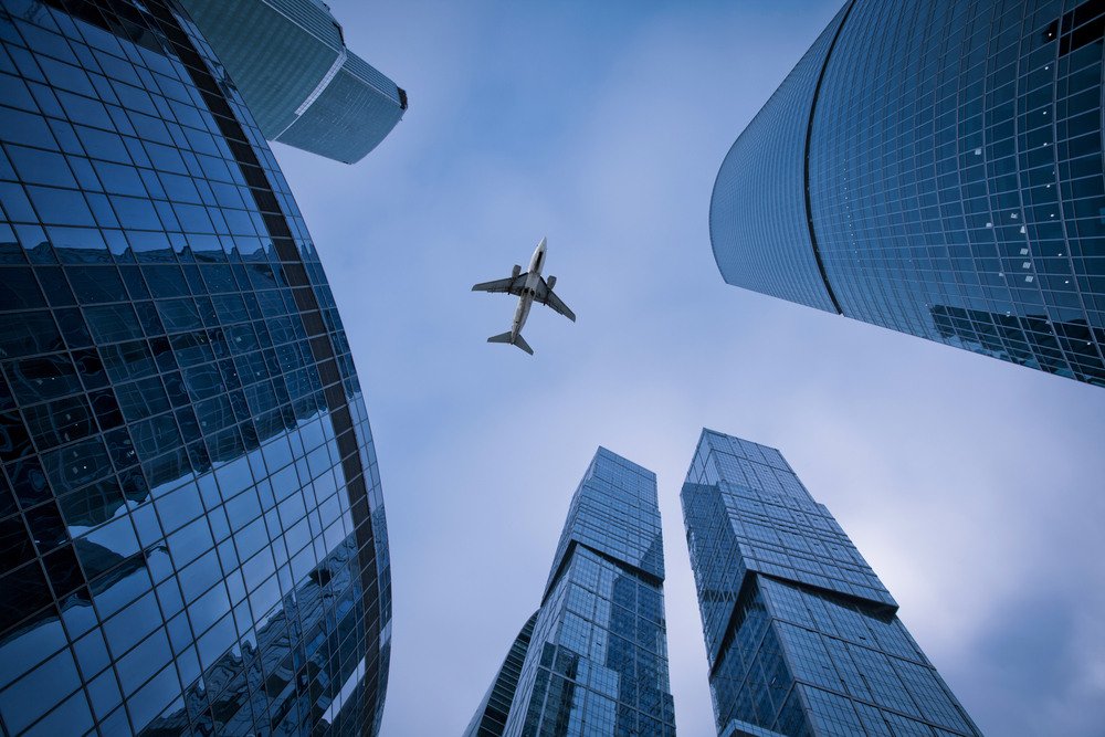 Photo of an airplane above the glass office buildings class="wp-image-167733" 