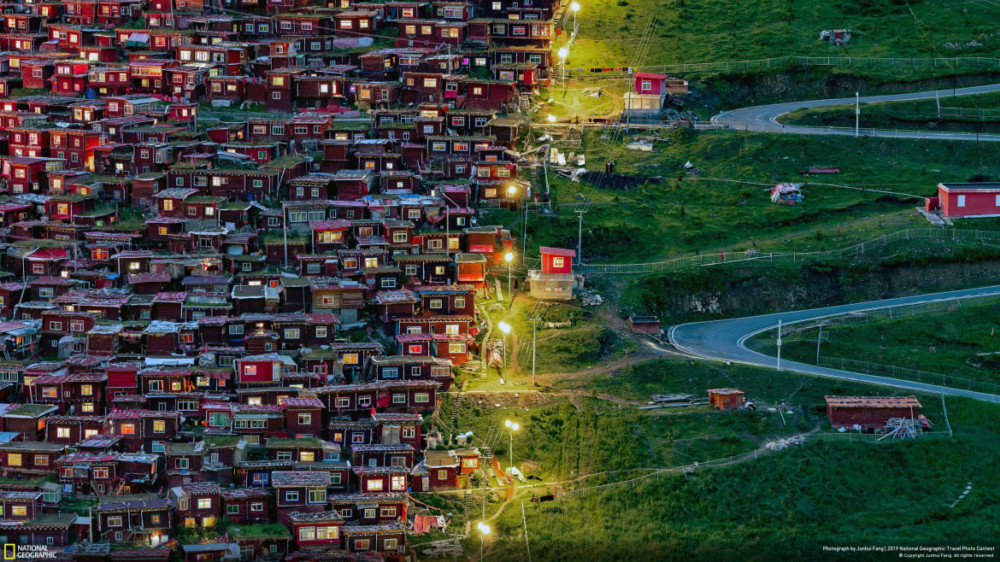Fot. Junhui Fang, &quot;FOLLOW THE LIGHT&quot; - nagroda publiczności w kategorii &quot;Cities&quot;. Zdjęcie zostało zrobione w Akademii Buddyjskiej Seda Larung Gar. Dojazd do Larung Gar z najbliższego miasta zajmuje około 14 godzin, a podróż jest dość trudna z powodu górskich dróg. Widok pokazuje małe czerwone domy po lewej stronie i puste zielone drogi po drugiej stronie. Mnisi podążają za światłami, aby wrócić do domu.  class="wp-image-956672" 